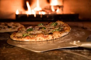 Pizza artesanal sobre uma pá metálica sendo colocada ou retirada do forno a lenha com chamas visíveis ao fundo, na Pizzaria Bellissima Italia em Luís Correia.