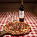 Cena de jantar italiano na pizzaria e restaurante Bellissima Italia: pizza artesanal com diversos ingredientes, acompanhada de uma garrafa de vinho tinto e duas taças, em ambiente acolhedor.