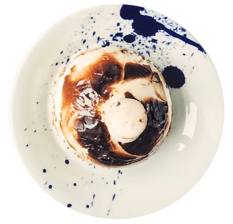 Sobremesa individual redonda com topo dourado/caramelizado e um detalhe cremoso no centro, servida em prato branco com decoração artística de calda azul, no Restaurante Bellissima Italia em Luís Correia.