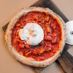 Pizza artesanal com molho de tomate vermelho intenso, tomates cereja e uma grande bola de queijo branco fresco (burrata ou muçarela de búfala) no centro, servida na Pizzaria Bellissima Italia em Luís Correia.